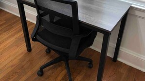 A modern office chair with wheels, positioned in front of a wooden desk with a metal frame, against a wall with a window with blinds.