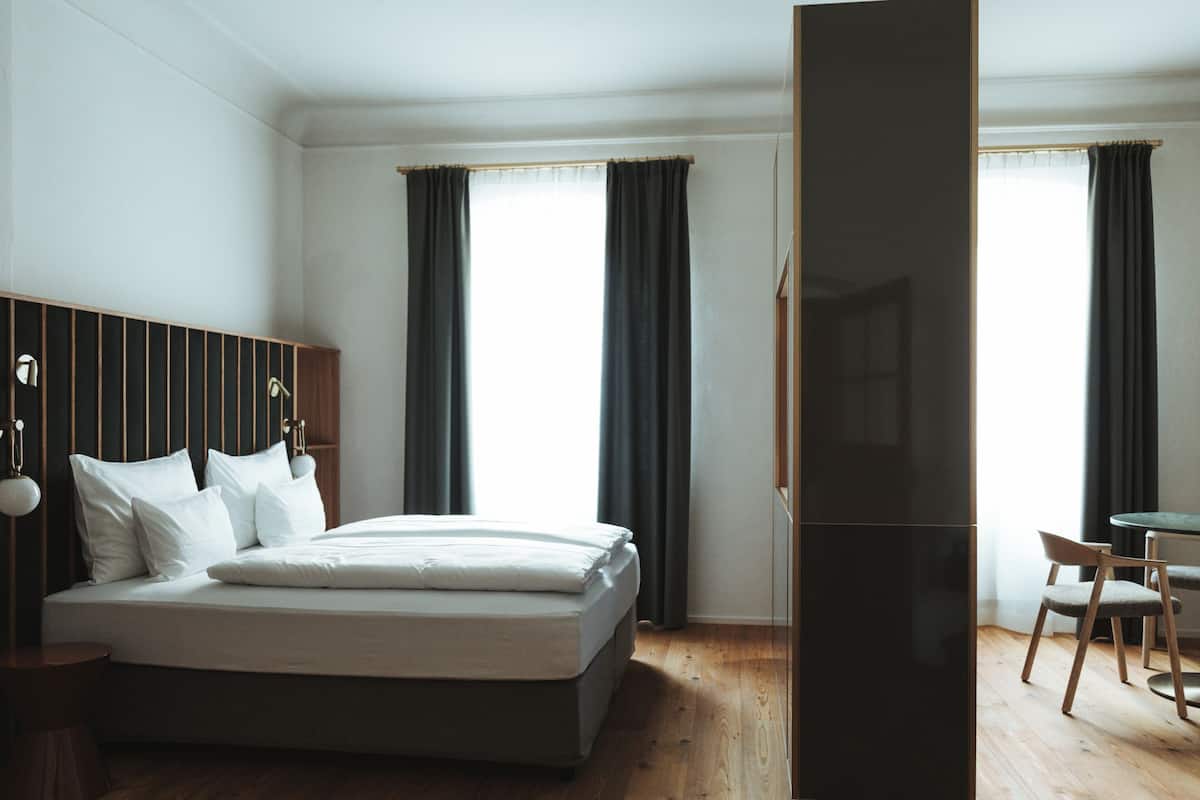 Ein modernes Hotelzimmer mit einem großen Bett, einem Holzkopfschild, einem Schreibtisch mit Stuhl und Blick auf die Stadt durch große Fenster.