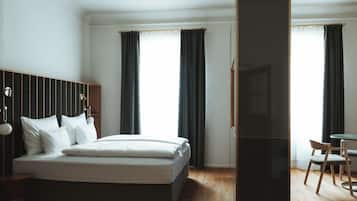 A modern hotel room with a large bed, a wooden headboard, a desk with a chair, and a view of the city through large windows.