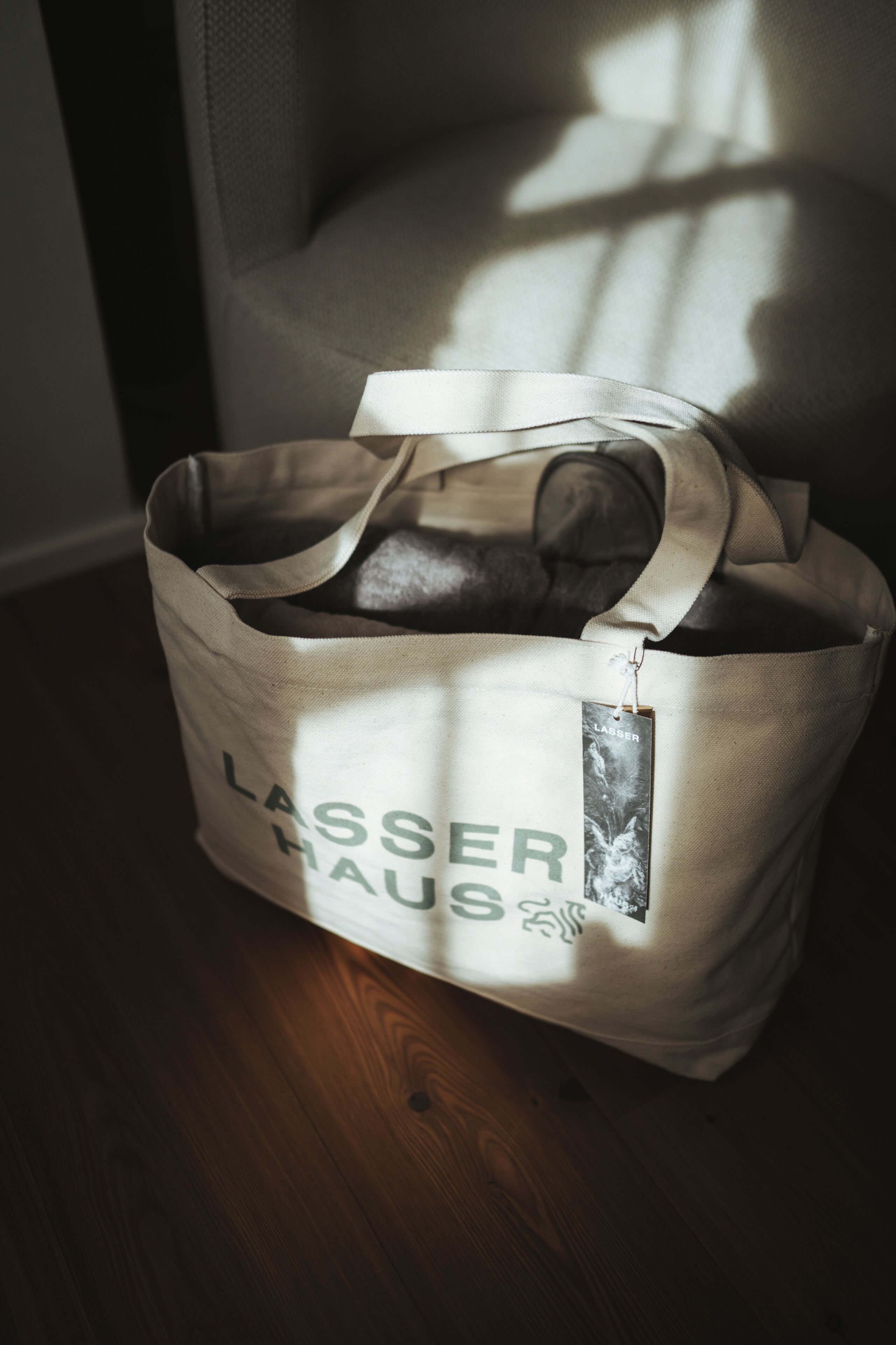 Una borsa porta tutto in tela beige con la scritta "LASSER HAUS", posata su un pavimento di legno.