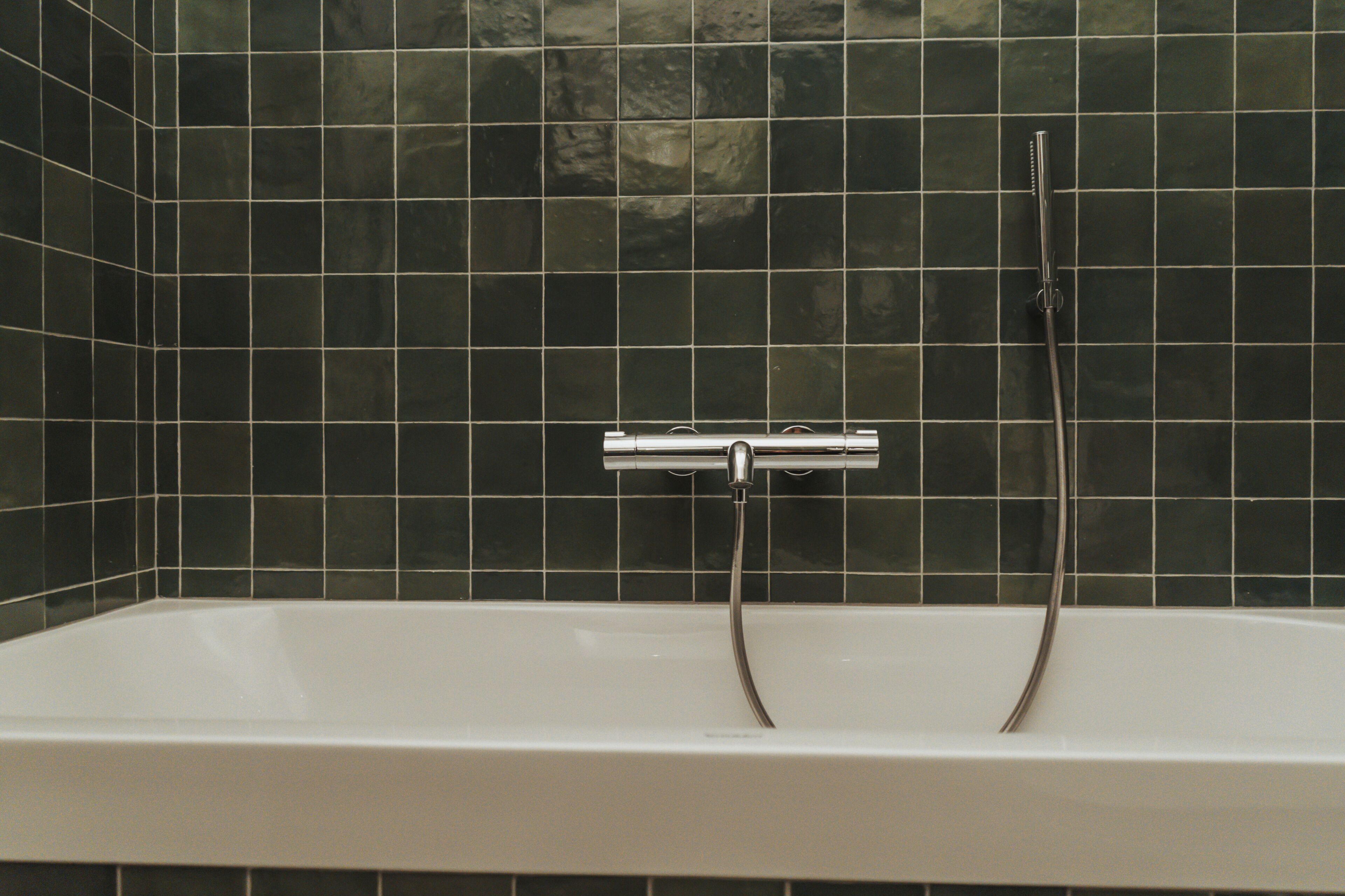 A bathroom with a white bathtub, dark green tiled walls, and a chrome faucet.