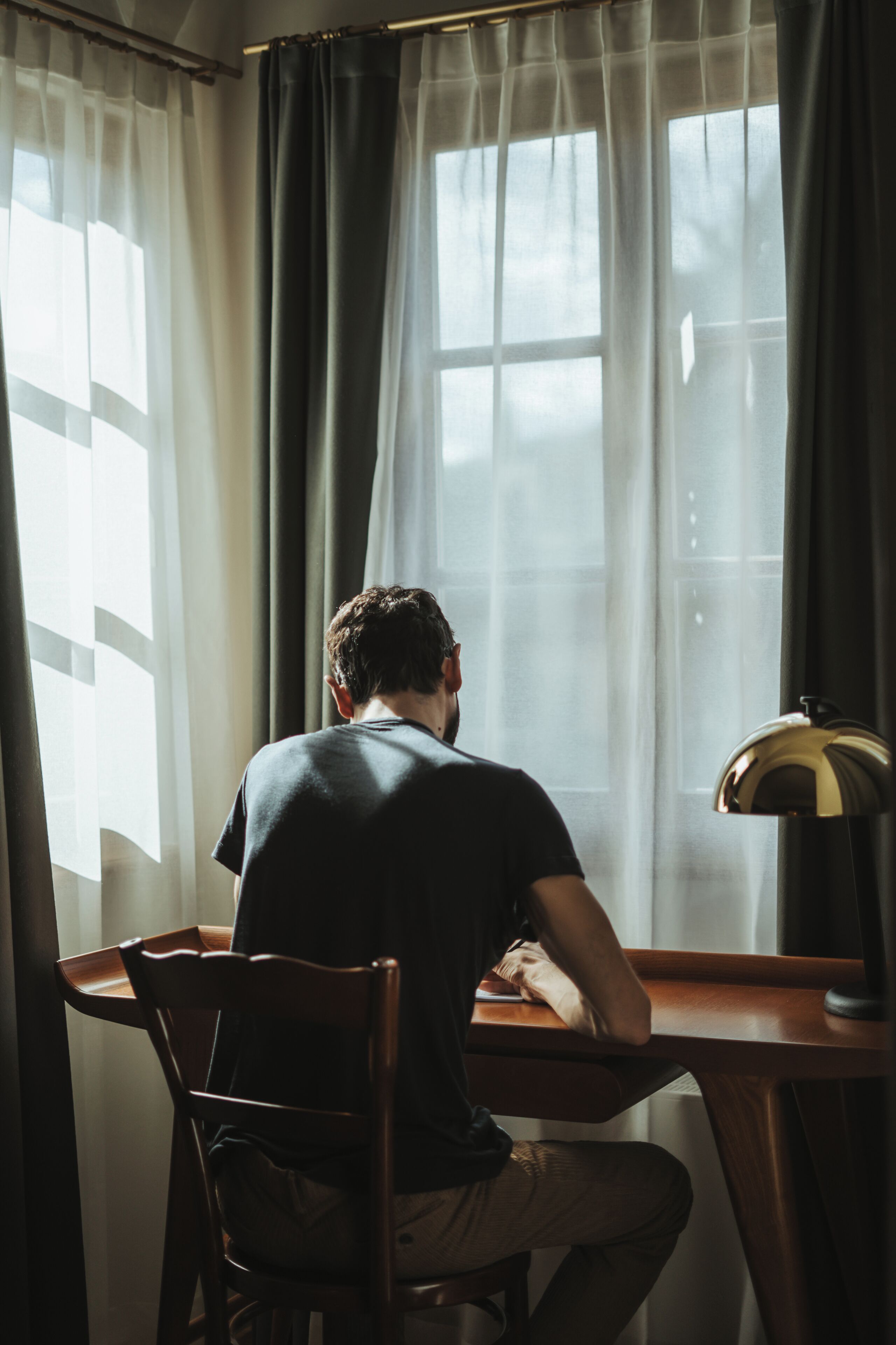 Une personne est assise à un bureau en bois, avec une lampe, près d’une fenêtre ornée de rideaux légers.
