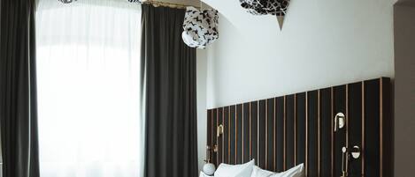 Habitación de hotel con cama, cortinas, cabecera con líneas verticales y lámparas colgantes.