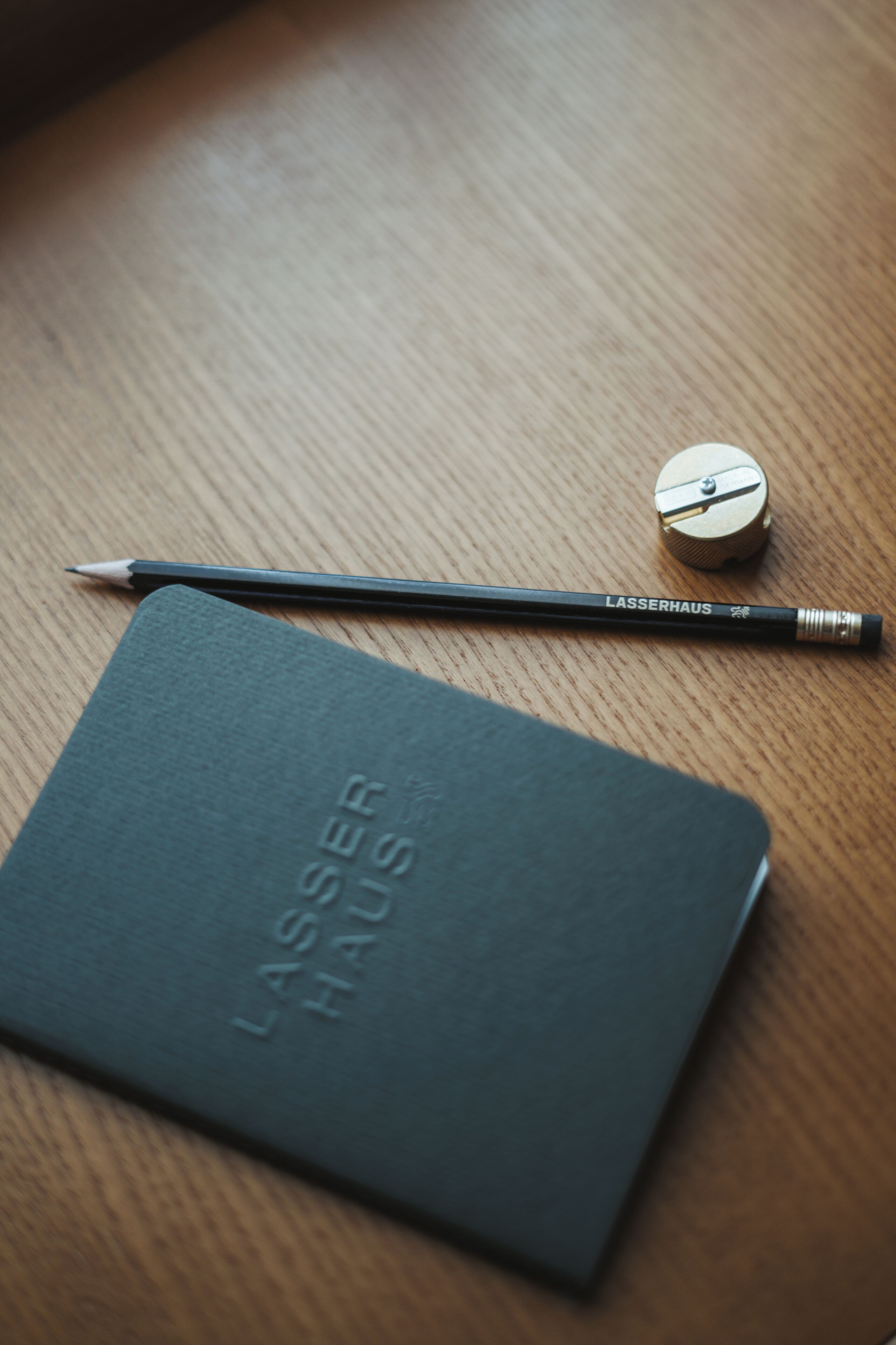 A dark green notebook with "LASSERHAUS" embossed on the cover, a black pencil, and a metallic pencil sharpener on a wooden surface.