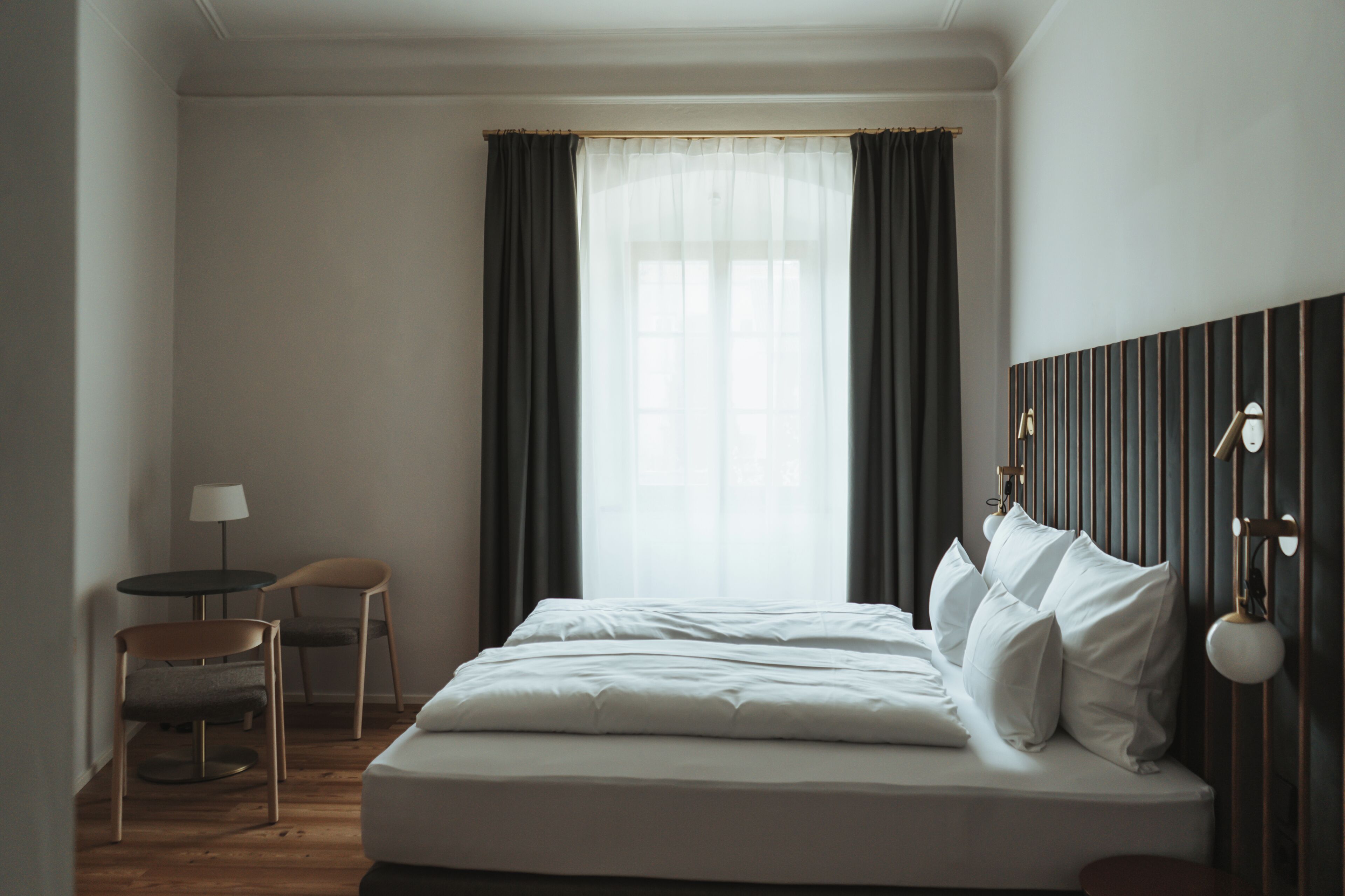 Een hotelkamer met een bed, twee stoelen, een lamp en een raam met gordijnen.