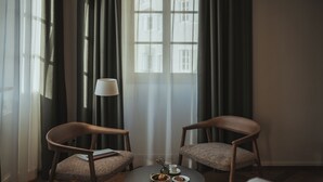 Una habitación con dos sillas de madera, una mesita redonda con una lámpara, una ventana con cortinas y una cama con ropa de cama blanca.