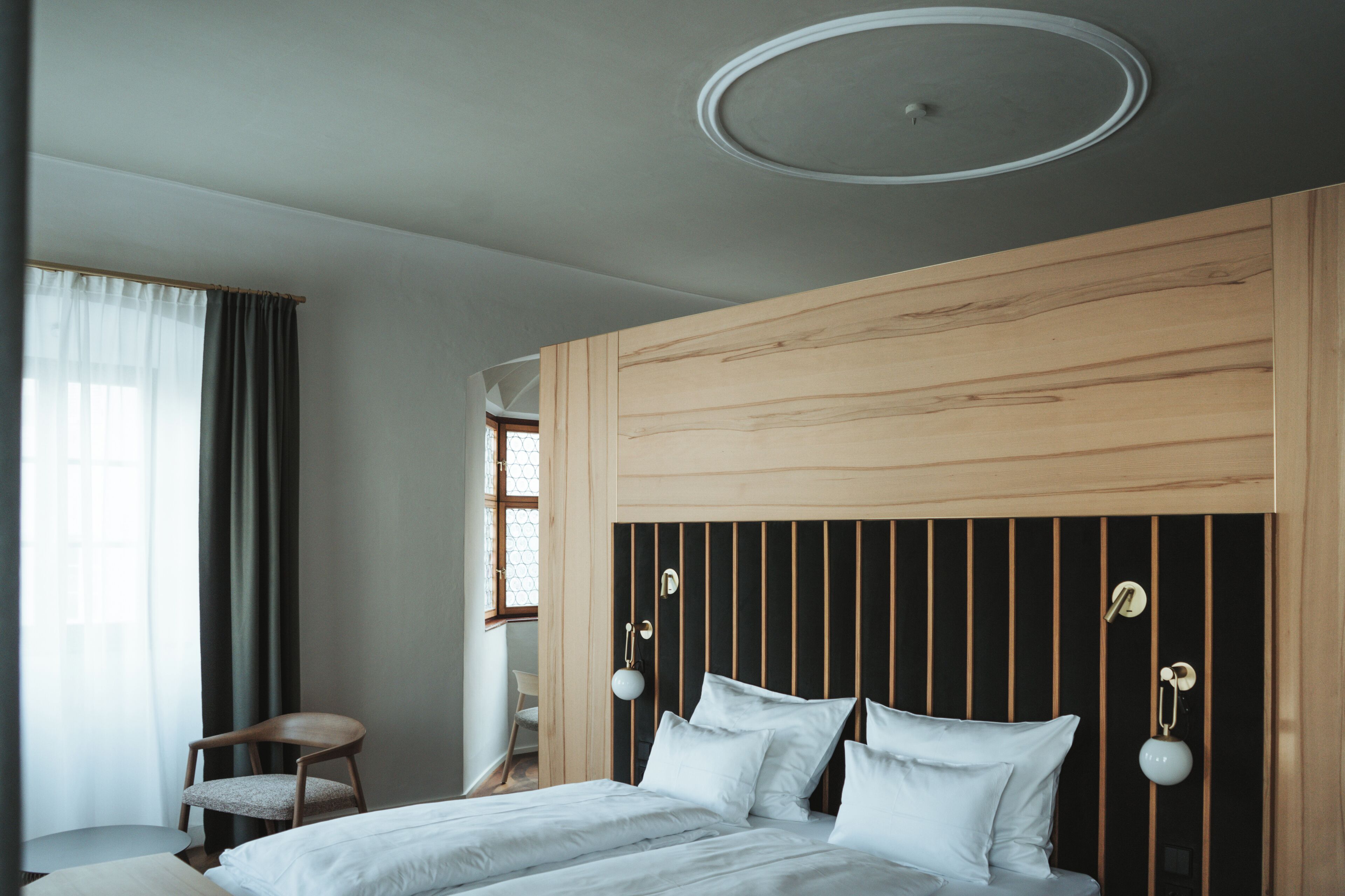 Una habitación de hotel moderna con un cabecero de madera, una cama con sábanas blancas, una silla y una ventana con cortinas.
