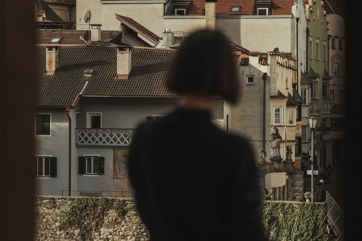 Eine Person steht auf einem Balkon und blickt auf eine Stadt mit historischen Gebäuden und zwei markanten Türmen.