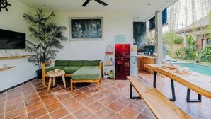 Un espacio de estar con sofá, una mesita, una banca, ventilador de techo, televisión y piscina visible a través de la ventana.