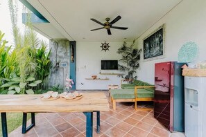 Una habitación con ventilador de techo, televisión, cama, mesa con comida y refrigerador.