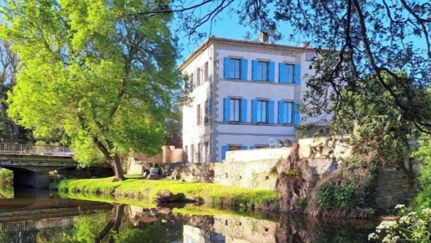 Un edificio reflejado en el agua, rodeado de vegetación y un puente de piedra.