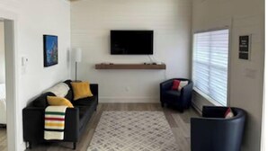 Ein modernes Wohnzimmer mit Holzboden, einem Wand-Fernseher, einer Couch mit gestreifter Tagesdecke und einem gemusterten Teppich.