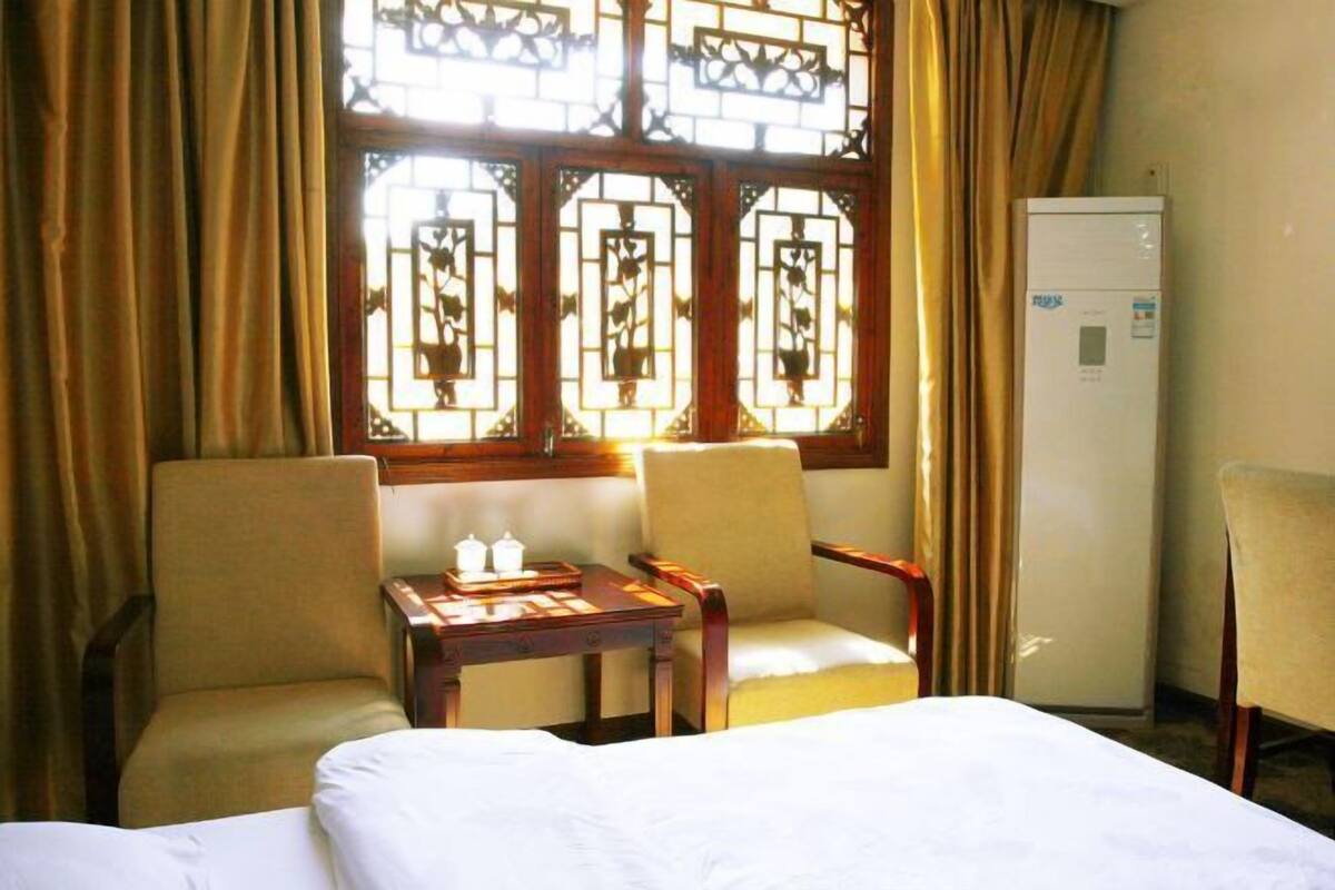 Une chambre d’hôtel comprenant un lit, deux chaises, une petite table avec un service à thé, un réfrigérateur et une fenêtre ornée d’un treillis décoratif.