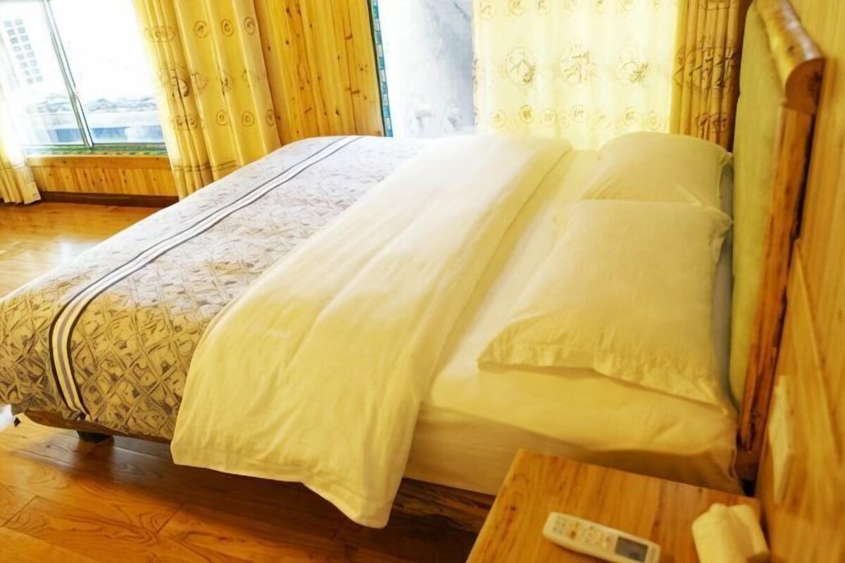 A bed with a patterned bedspread, a white blanket, and pillows. There is a wooden nightstand with a remote control and a tissue box.