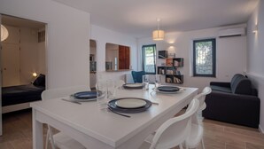 A modern, well-lit room with a dining table set for four, a sofa, a bookshelf, and a window with a view.