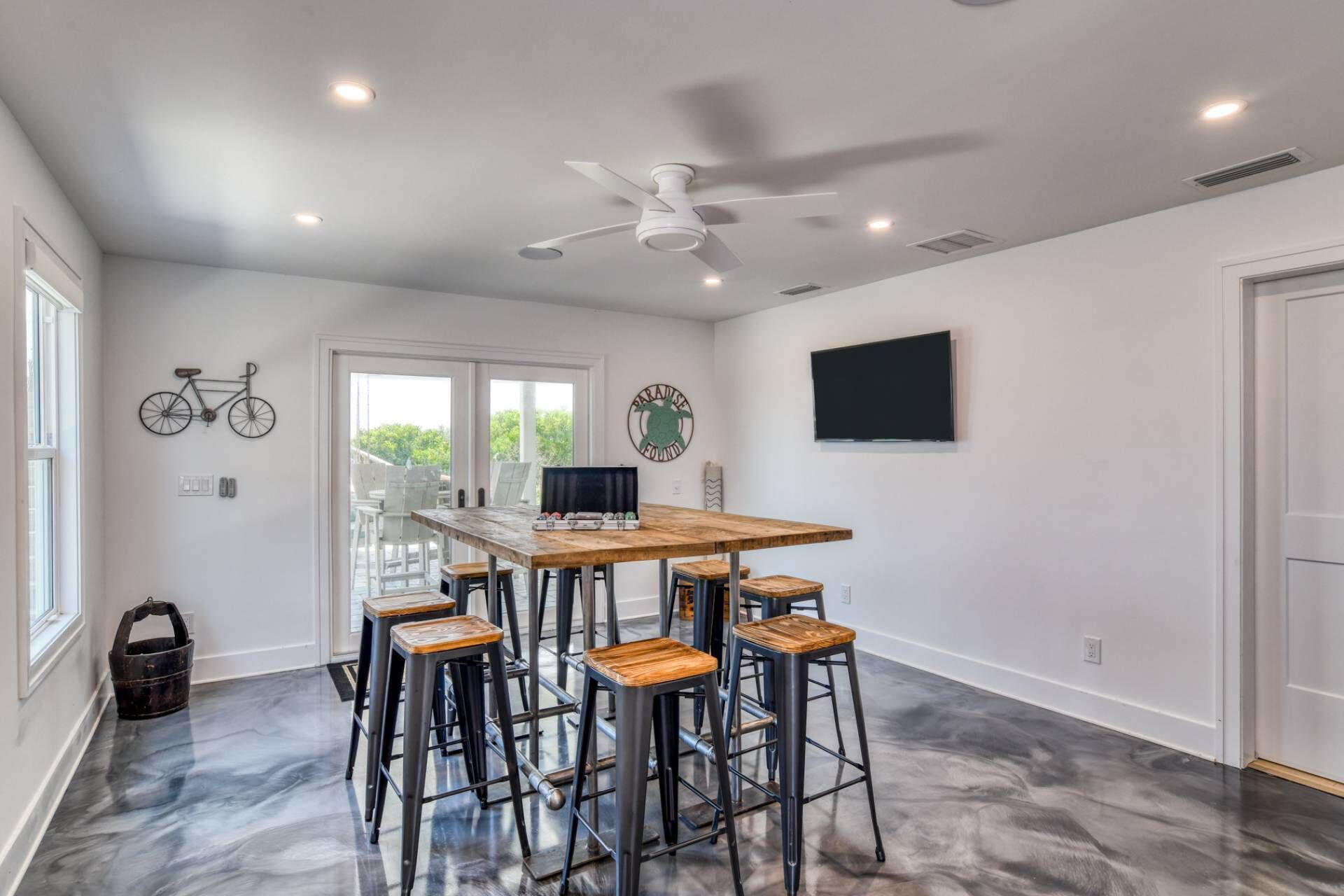 Una habitación amplia con una mesa de madera y taburetes de bar, un ventilador de techo y un televisor montado.