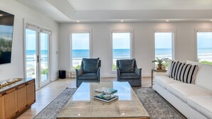 Ein geräumiges Wohnzimmer mit einem großen Glastisch, zwei Sesseln, einer Couch und Meerblick durch große Fenster.
