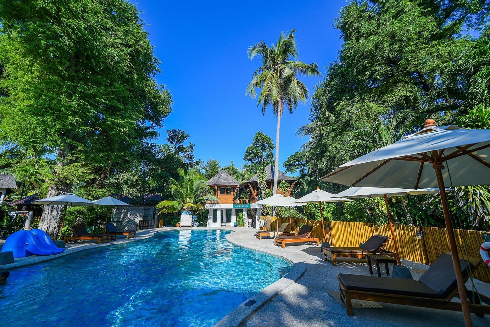 A swimming pool surrounded by lounge chairs and umbrellas, with a tropical setting and wooden structures in the background.