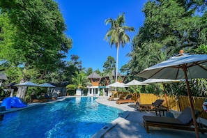 Una piscina rodeada de sillas de descanso y sombrillas, en un entorno tropical y con estructuras de madera al fondo.