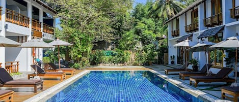 A pool area with lounge chairs and umbrellas, surrounded by lush greenery and a building with multiple balconies.
