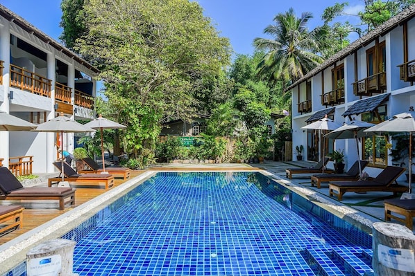 Un área de piscina con sillones de descanso y sombrillas, rodeada de abundante vegetación y un edificio con varios balcones.