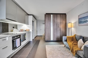 A modern kitchen with a central island, built-in appliances, and a sliding door leading to a living area with a sofa and a lamp. - Chic Urban Studio Cloud Nine - 1B1B (Melbourne)