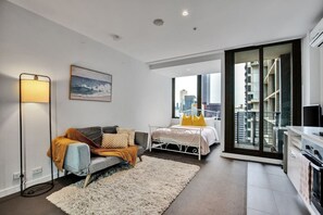 Quarto moderno com cama, sofá, luminária de chão e vista da cidade através de janelas amplas.
