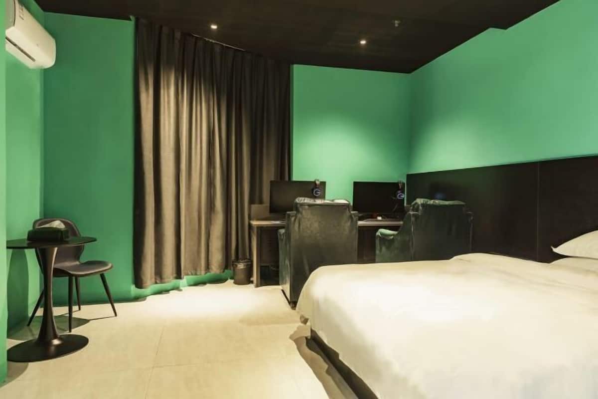 Quarto de hotel com parede verde, uma cama, uma escrivaninha com um computador e uma cadeira.