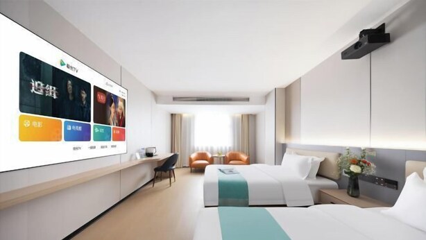 A hotel room with a bed, a desk, a chair, a TV screen displaying content, and a window with curtains.