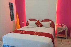 Un lit avec une literie blanche et un plaid rouge, une table de chevet avec une lampe, et une fenêtre donnant sur l’extérieur.