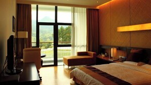 A hotel room with a bed, a desk, a chair, and a large window with a view of greenery.