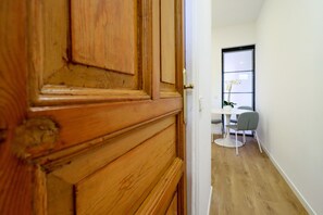 A wooden door leading to a room with a small table and a chair, a window with blinds, and a plant. - ChicLavapies Center (Madrid)