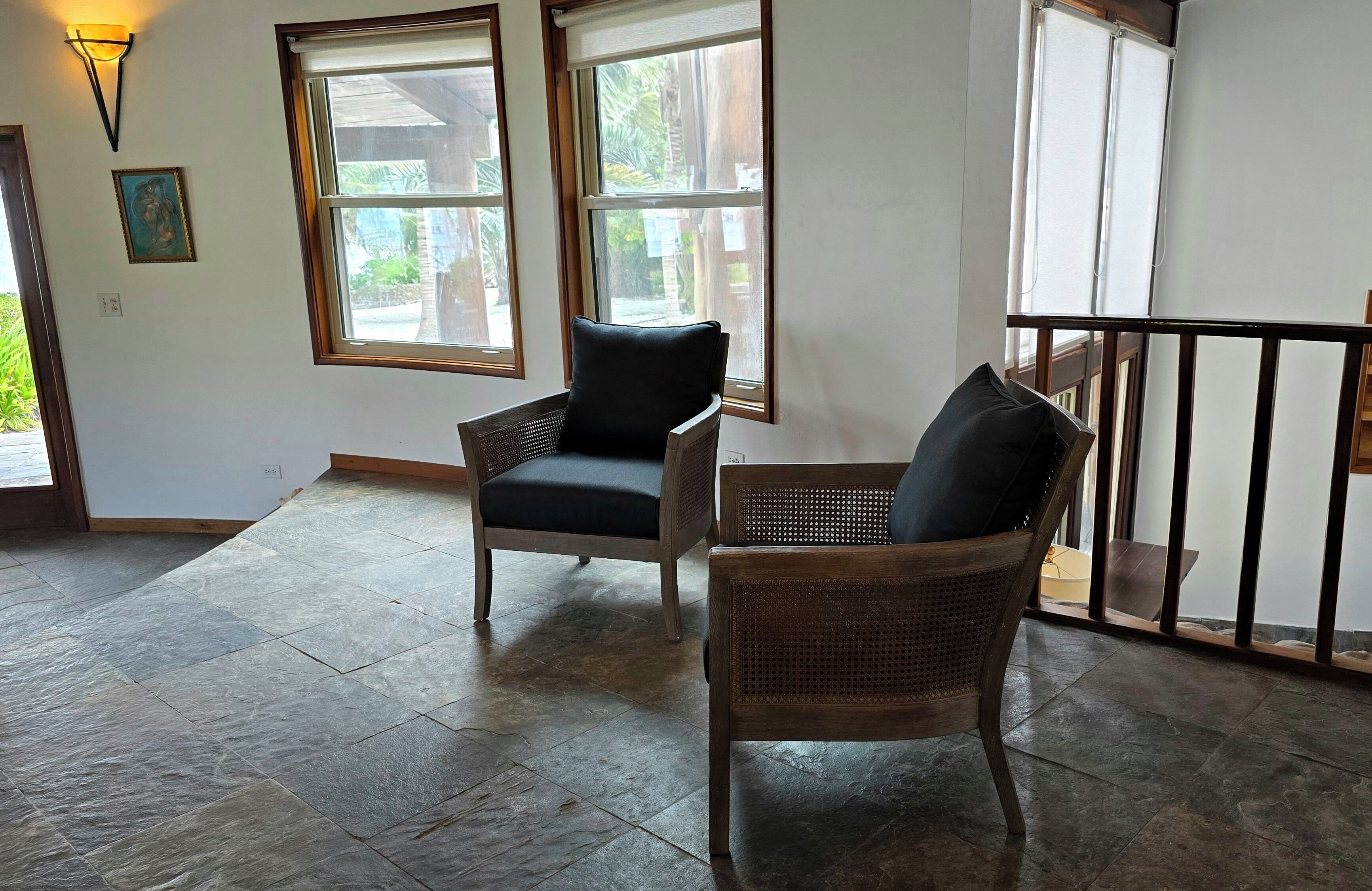A room with two wicker chairs, large windows, a stone floor, and a balcony railing.