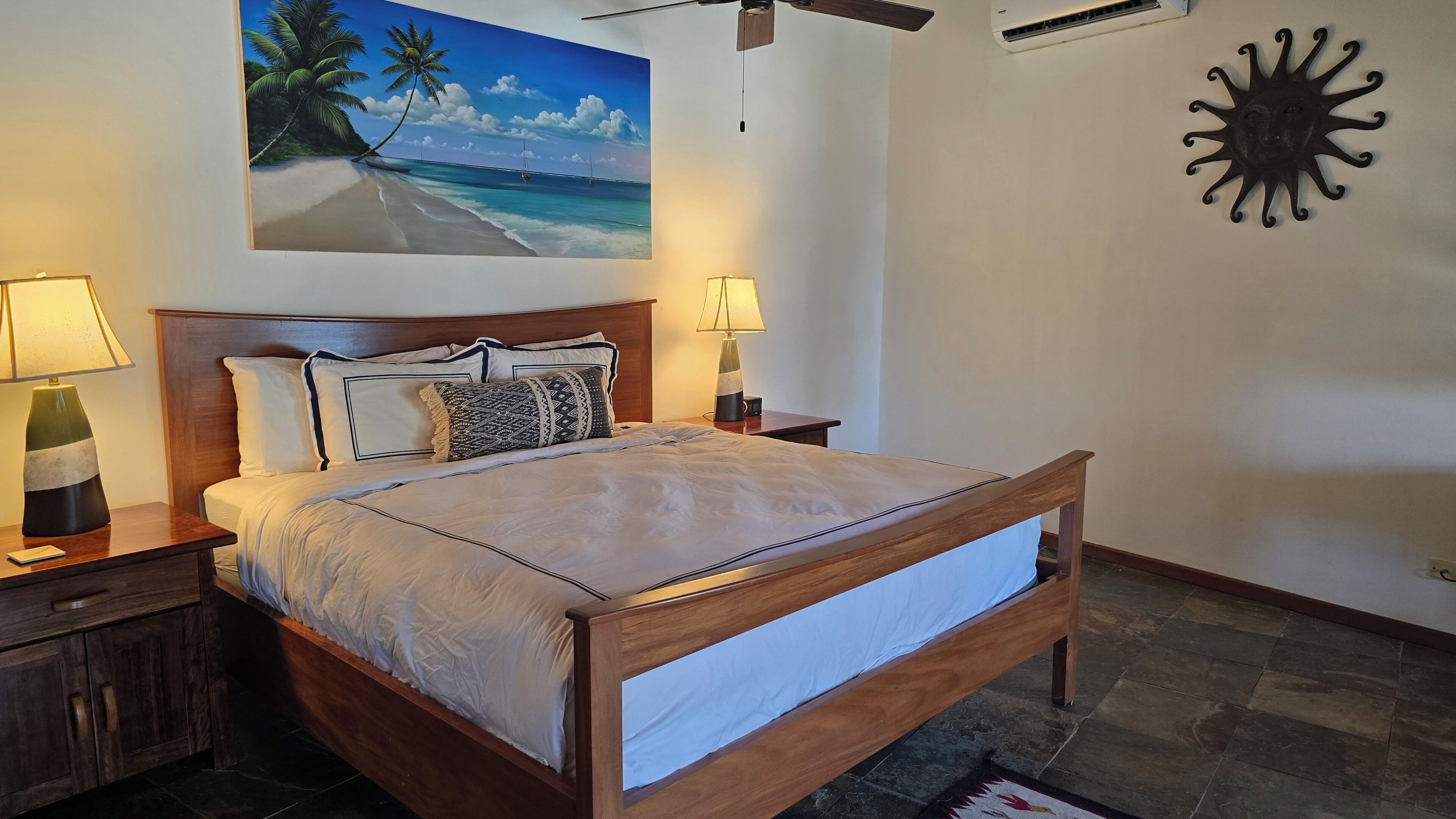 A bedroom with a bed, nightstands, a painting of a beach, a wall clock, and a ceiling fan.