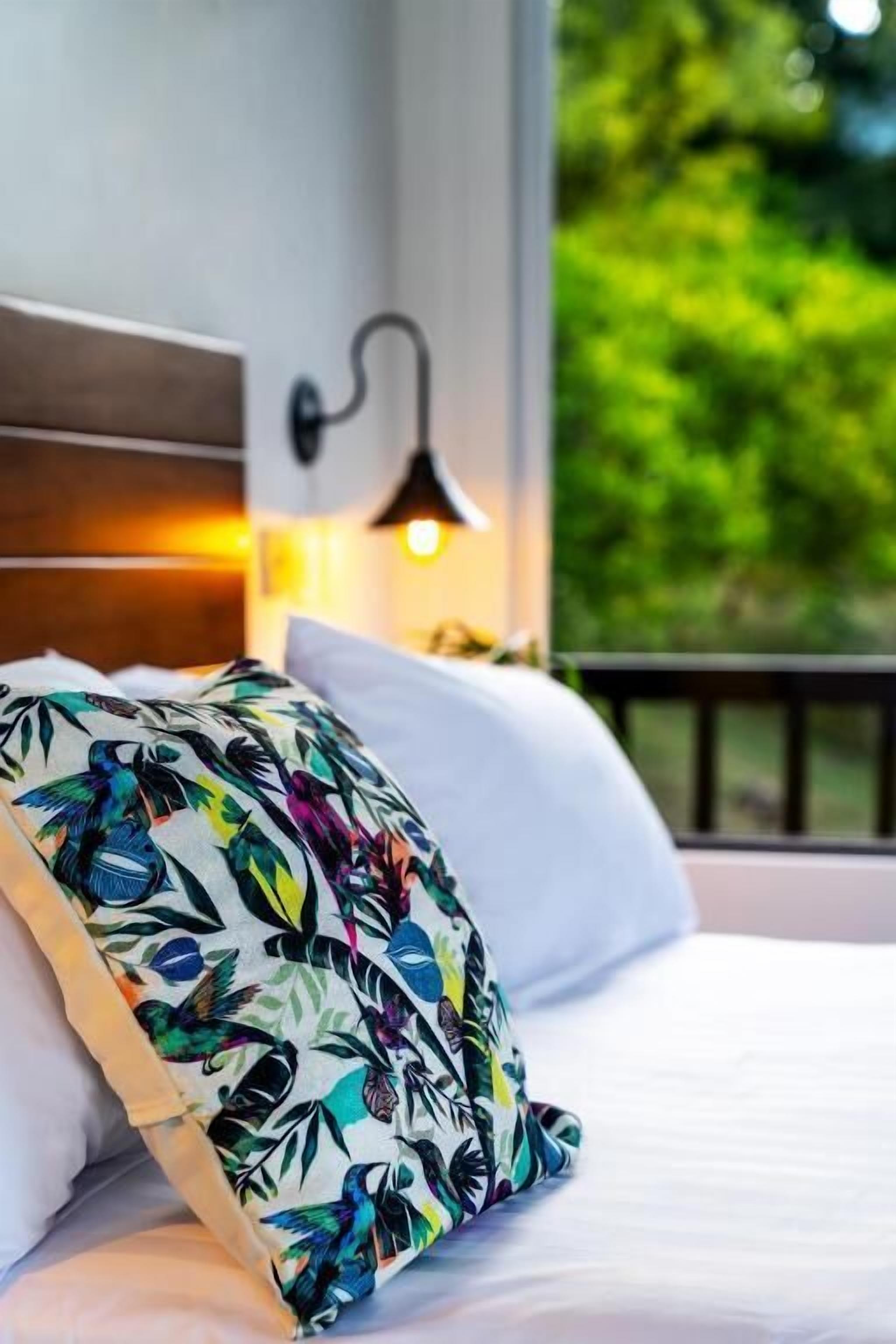 Una cama con sábanas blancas y un cojín de colores con estampado de aves tropicales.