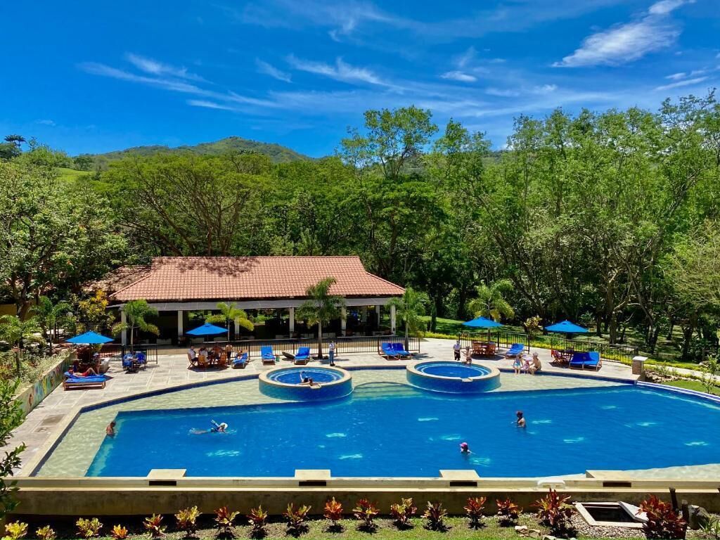 Una piscina con varias pistas, rodeada de sillas de descanso y sombrillas, con un edificio al fondo.