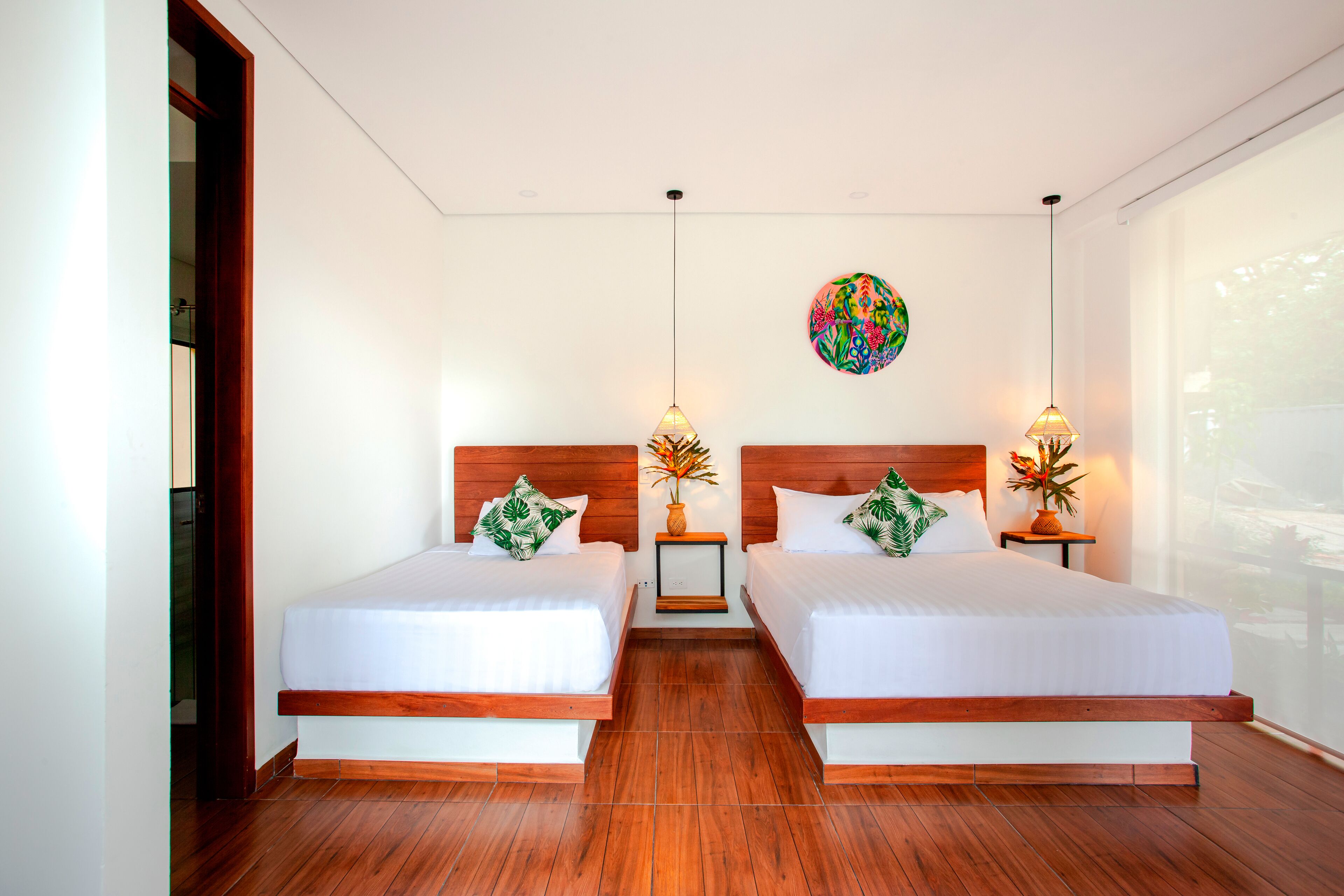 A hotel room with two beds, wooden headboards, white bedding, small side tables with lamps, and a large window with a view.
