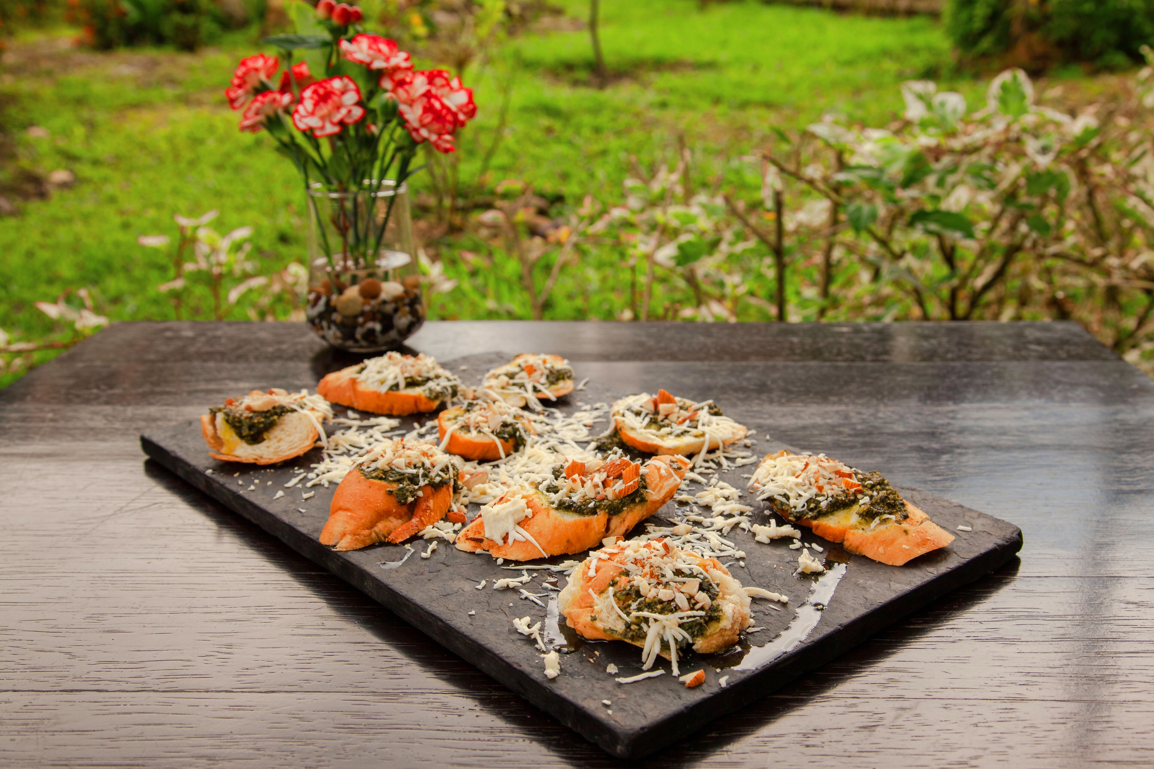 A slate serving board with appetizers, a vase of red flowers, and a garden background.