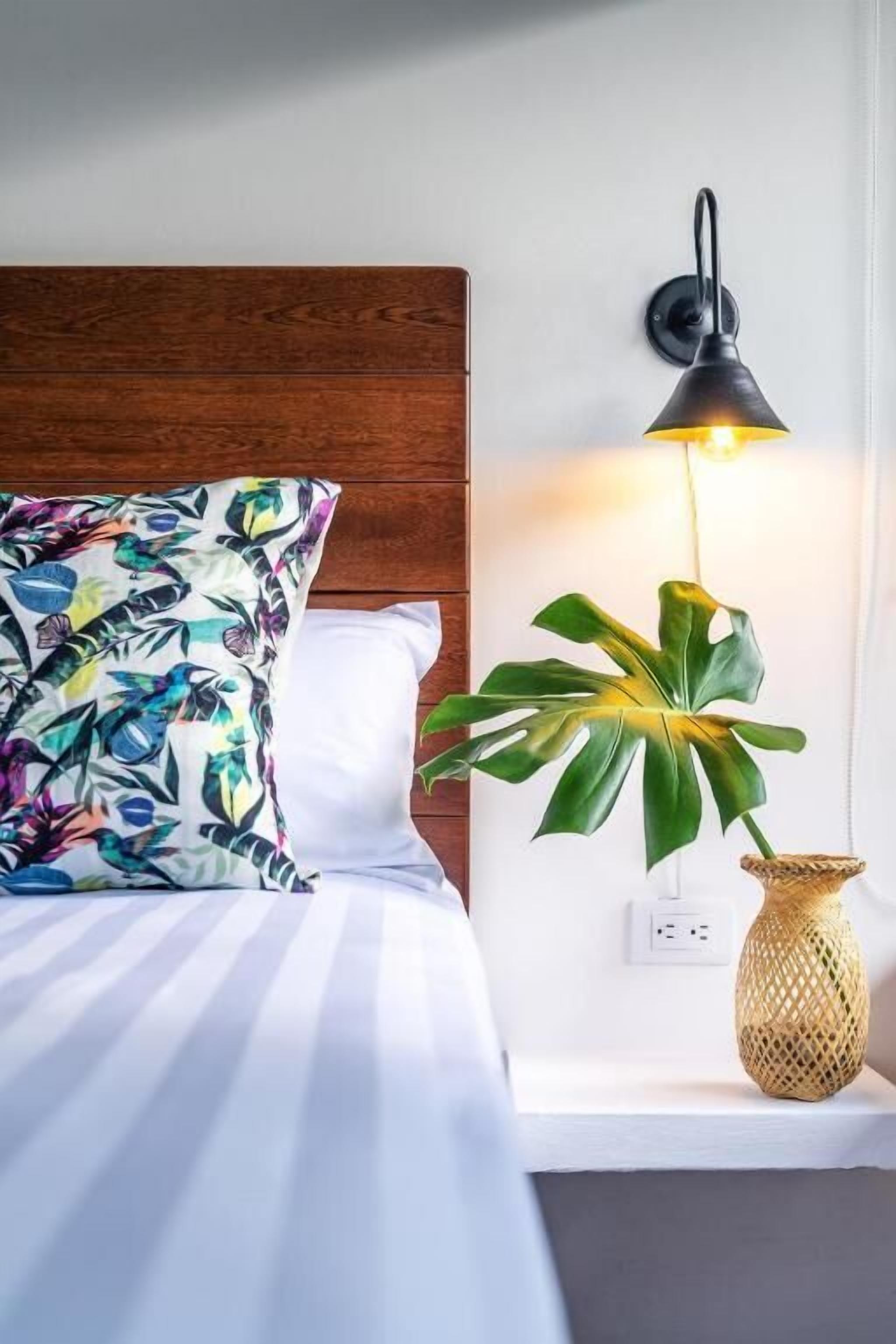 A neatly made bed with a colorful floral pillow and a white pillow, a wooden headboard, a wall-mounted lamp, and a large green plant in a woven pot.