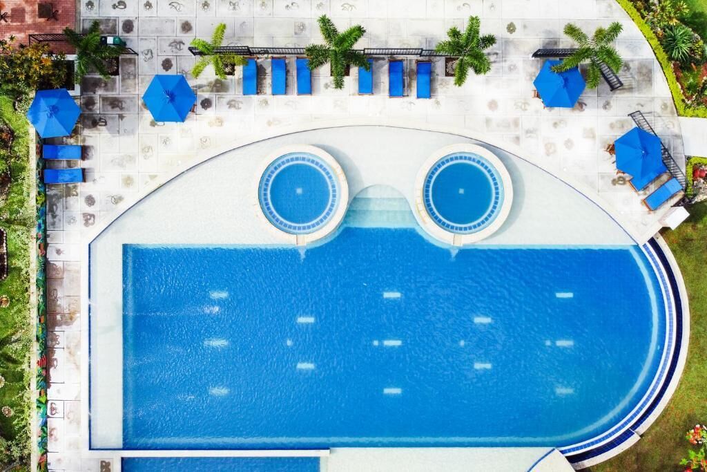 A large outdoor swimming pool with two circular hot tubs, surrounded by lounge chairs and umbrellas.