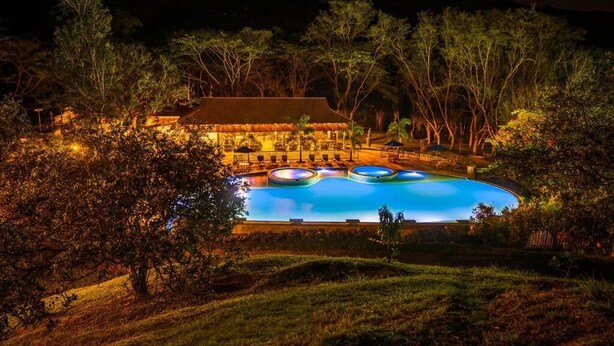 A nighttime view of a resort with a large swimming pool surrounded by trees and a building with a thatched roof. - Hotel Nana  (Villeta)