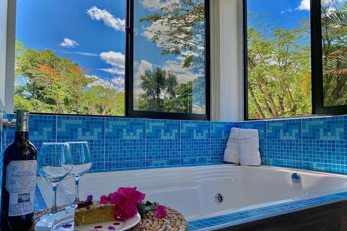 A bathroom with a bathtub, a bottle of wine, two glasses, and a plate with a slice of cake and flowers.