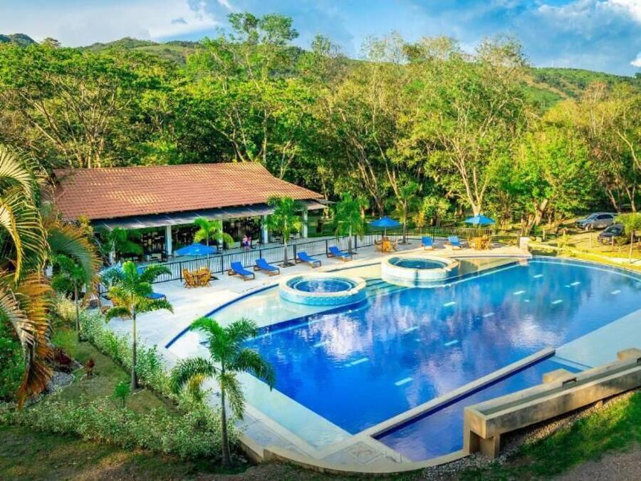 Una amplia piscina al aire libre rodeada de exuberante vegetación, con un área de descanso cubierta y un edificio pequeño al fondo.