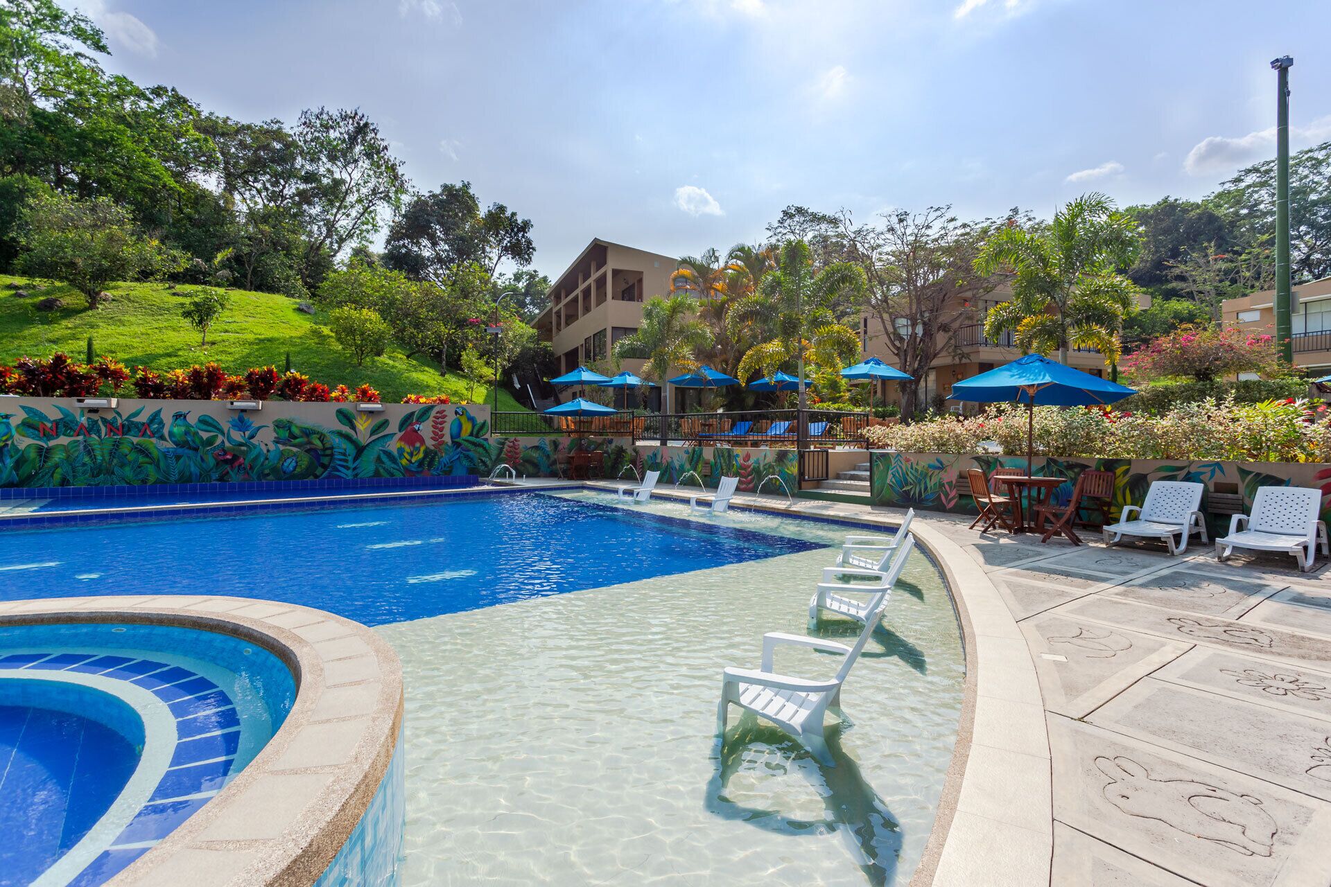 Ein Swimmingpool mit klarem Wasser, umgeben von Liegestühlen und Sonnenschirmen. Im Hintergrund ist eine Wandmalerei und viel Grünwerk zu sehen.