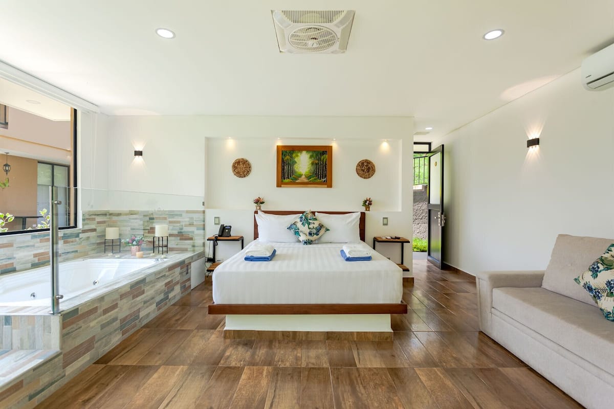 A modern hotel room with a large bed, a glass-enclosed balcony, a jacuzzi, and a view of the outdoors.
