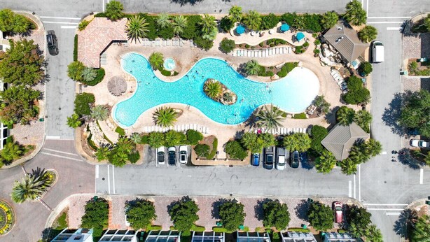 Vista aérea de un complejo turístico con una piscina central, rodeado de palmeras y un estacionamiento.