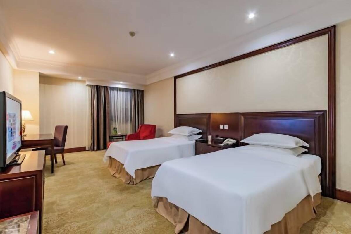 Ein Hotelzimmer mit zwei Betten, einem Schreibtisch mit Stuhl, einem Fernseher und einem Fenster mit Vorhängen.