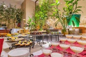 Montaje de bufé con diferentes alimentos y bebidas, rodeado de plantas de interior y una estructura decorativa verde.