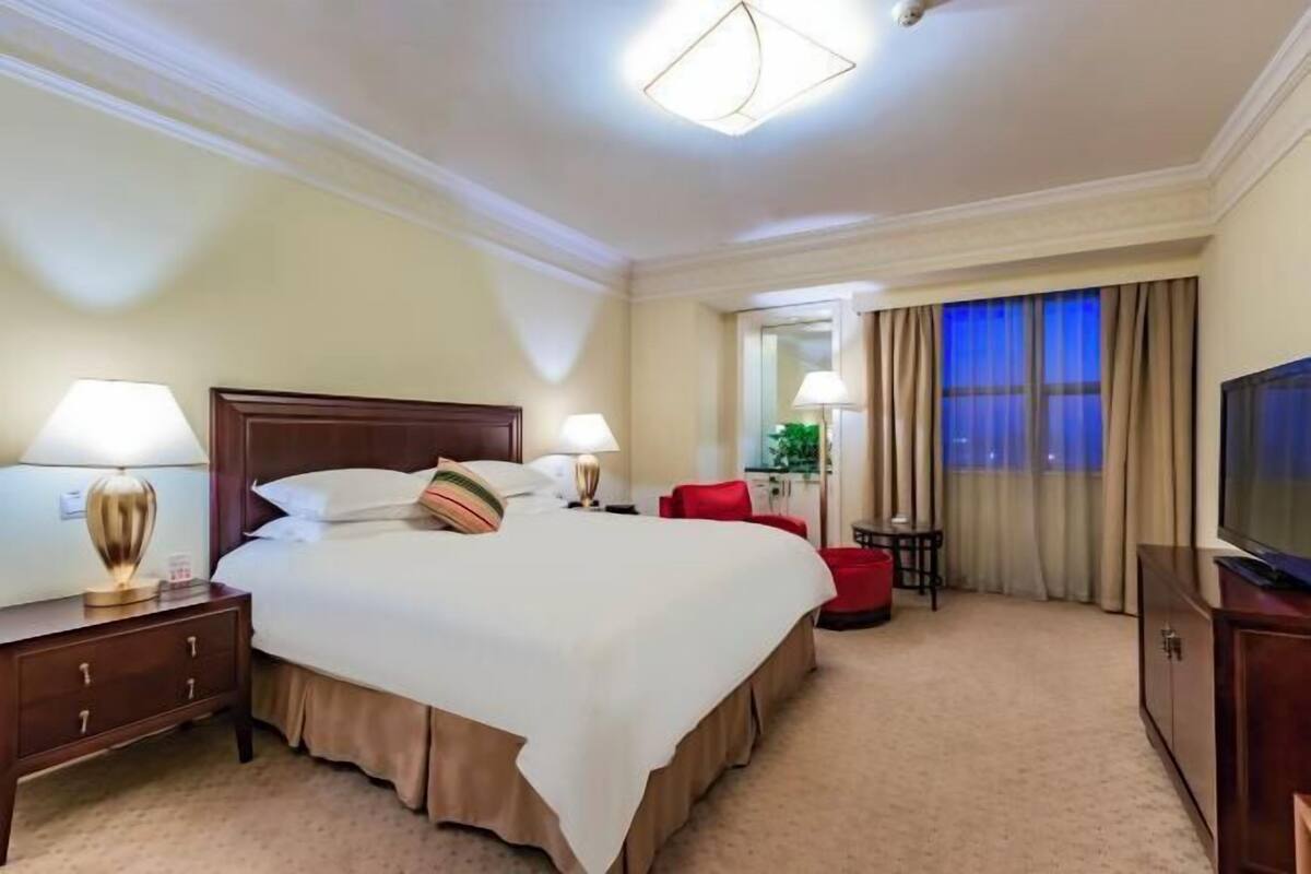 Ein Hotelzimmer mit einem großen Bett, zwei Nachttischlampen, einem Fernseher, einem Schreibtisch und einem Sessel.
