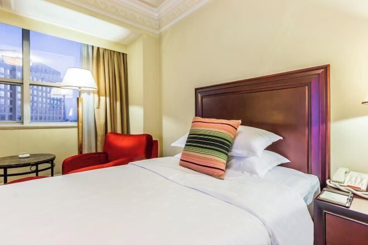 A hotel room with a large bed, a red chair, a small table, and a window with curtains.